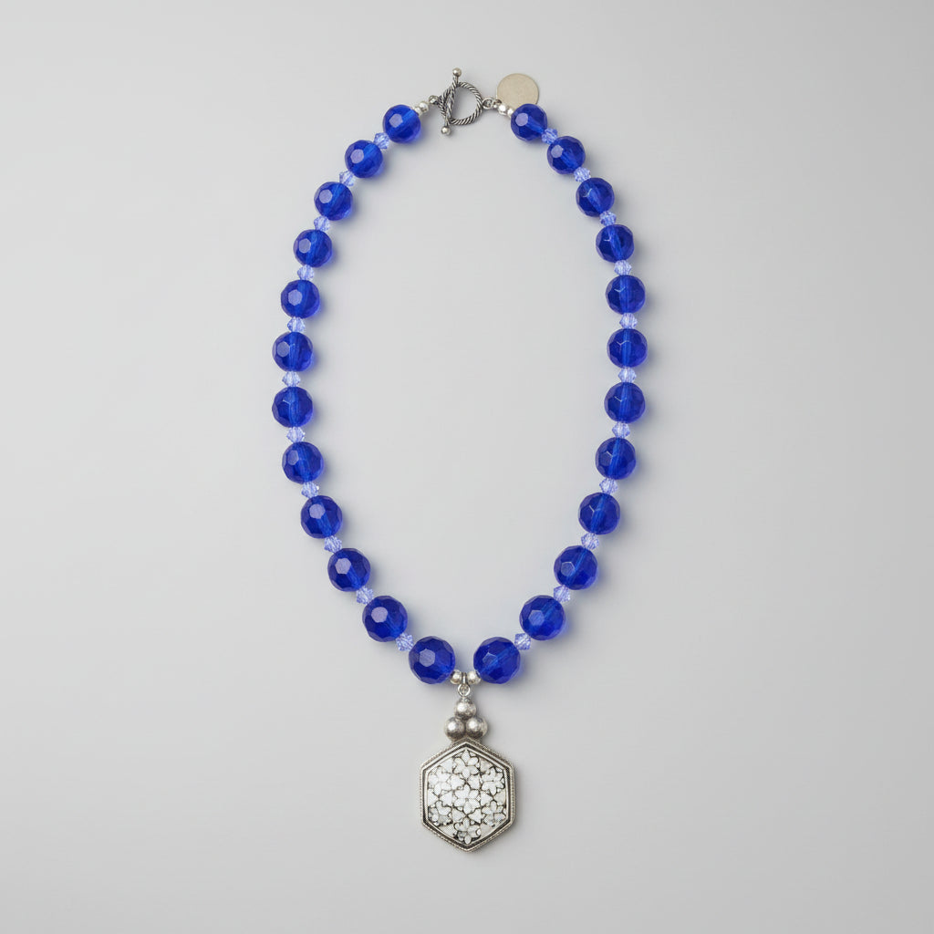 Brilliant cobalt blue takes center stage in this one-of-a-kind handcrafted Billielephante necklace. Rich, transparent faceted blue glass beads are individually separated by petite iridescent crystal-style spacers, creating exceptional sparkle as the necklace catches the light.

The strand leads to an oversized silver-tone geometric statement pendant, featuring an intricate mosaic-like reflective center and dimensional metal border. Decorative silver-tone hardware completes the eclectic artisan design.

Bold