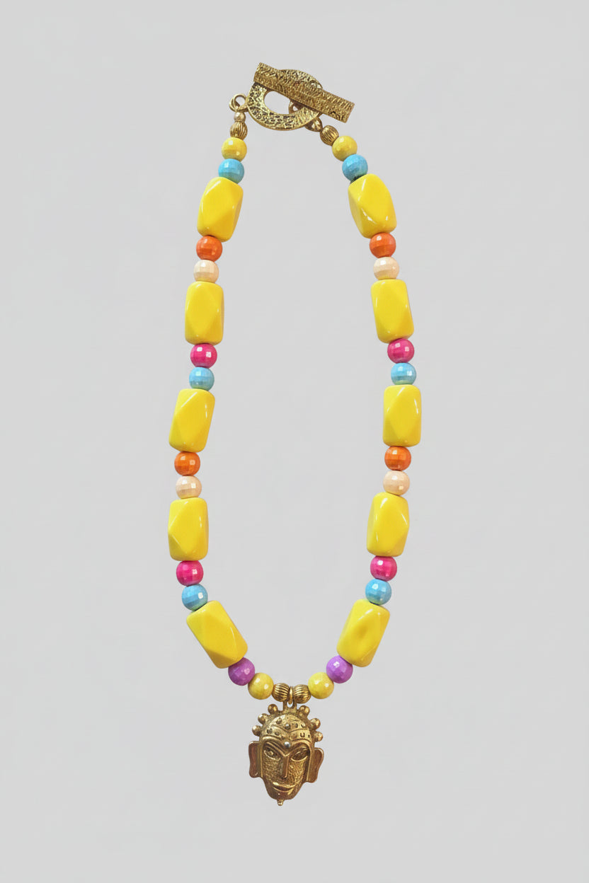 A bold, one-of-a-kind handcrafted necklace featuring oversized glossy yellow irregular beads accented with faceted beads in turquoise blue, coral-orange, fuchsia, cream and purple. Antique-gold-tone details add warmth throughout, while a sculptural mask-style pendant creates a dramatic focal point. Finished with an oversized textured antique-gold-tone toggle clasp, making even the closure a distinctive part of the design.