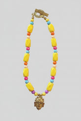 A bold, one-of-a-kind handcrafted necklace featuring oversized glossy yellow irregular beads accented with faceted beads in turquoise blue, coral-orange, fuchsia, cream and purple. Antique-gold-tone details add warmth throughout, while a sculptural mask-style pendant creates a dramatic focal point. Finished with an oversized textured antique-gold-tone toggle clasp, making even the closure a distinctive part of the design.