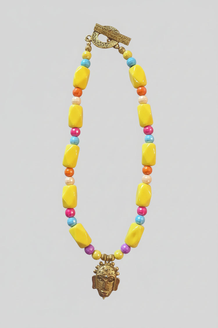 A bold, one-of-a-kind handcrafted necklace featuring oversized glossy yellow irregular beads accented with faceted beads in turquoise blue, coral-orange, fuchsia, cream and purple. Antique-gold-tone details add warmth throughout, while a sculptural mask-style pendant creates a dramatic focal point. Finished with an oversized textured antique-gold-tone toggle clasp, making even the closure a distinctive part of the design.