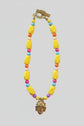 A bold, one-of-a-kind handcrafted necklace featuring oversized glossy yellow irregular beads accented with faceted beads in turquoise blue, coral-orange, fuchsia, cream and purple. Antique-gold-tone details add warmth throughout, while a sculptural mask-style pendant creates a dramatic focal point. Finished with an oversized textured antique-gold-tone toggle clasp, making even the closure a distinctive part of the design.